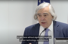 Video capture of Ernest Moniz speaking at a podium with the US flag behind him.